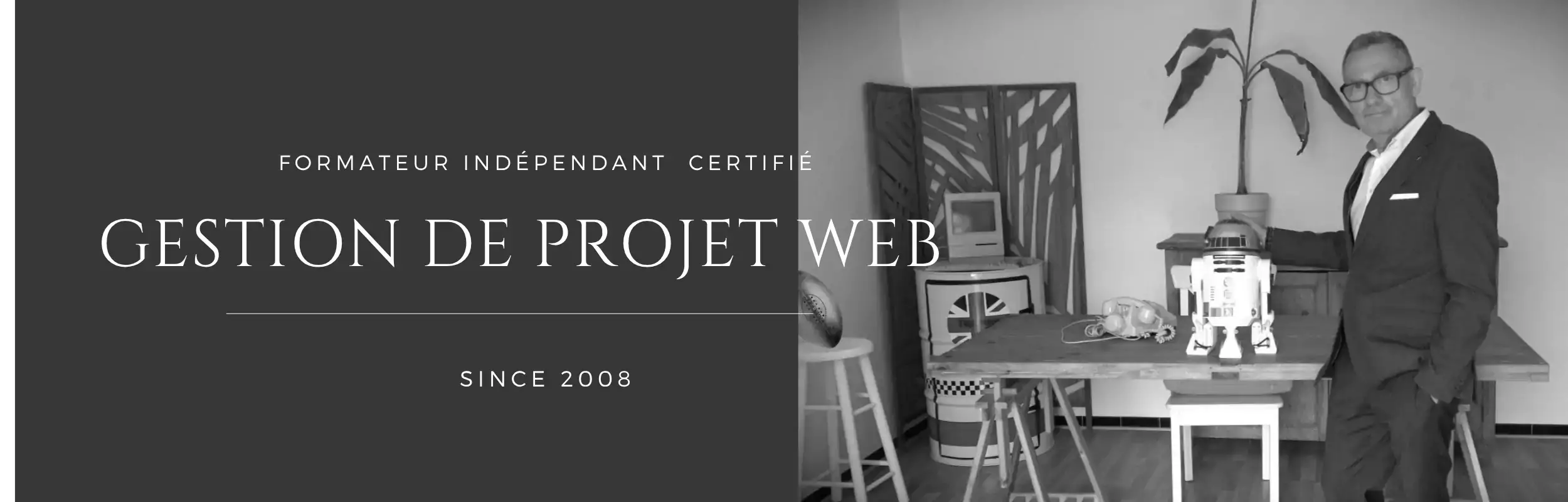 Photo en noir et blanc d'un homme en costume, formateur en Gestion de Projet Web, avec un robot sur une table en arrière-plan et le texte "Since 2008".